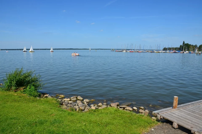 Ruhiger See mit Segelbooten und grünem Ufer im Hintergrund unter klarem, blauem Himmel.